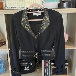 May include: Black jacket with gold-colored stud and rivet details. The jacket has a notched collar, a zip-up front, and a belt at the waist. The cuffs also feature decorative straps. The label reads "JOHN MURROUGH".
