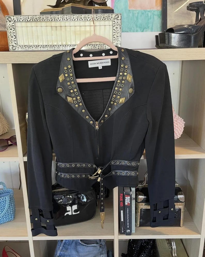May include: Black jacket with gold-colored stud and rivet details. The jacket has a notched collar, a zip-up front, and a belt at the waist. The cuffs also feature decorative straps. The label reads "JOHN MURROUGH".
