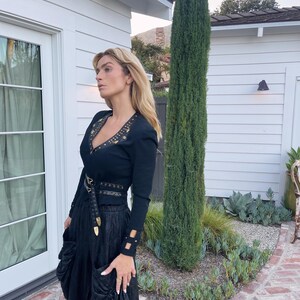 May include: A black outfit featuring a long-sleeved top with gold embellishments around the neckline and cuffs, paired with a high-waisted skirt and a belt with gold accents. The outfit is worn outdoors.
