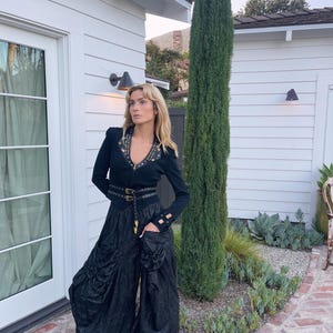 May include: A black long-sleeved top with gold-colored stud accents and a matching belt. The outfit includes wide-leg black pants and black heeled sandals. A tall, green tree stands in the background, with a white building and brick pathway.