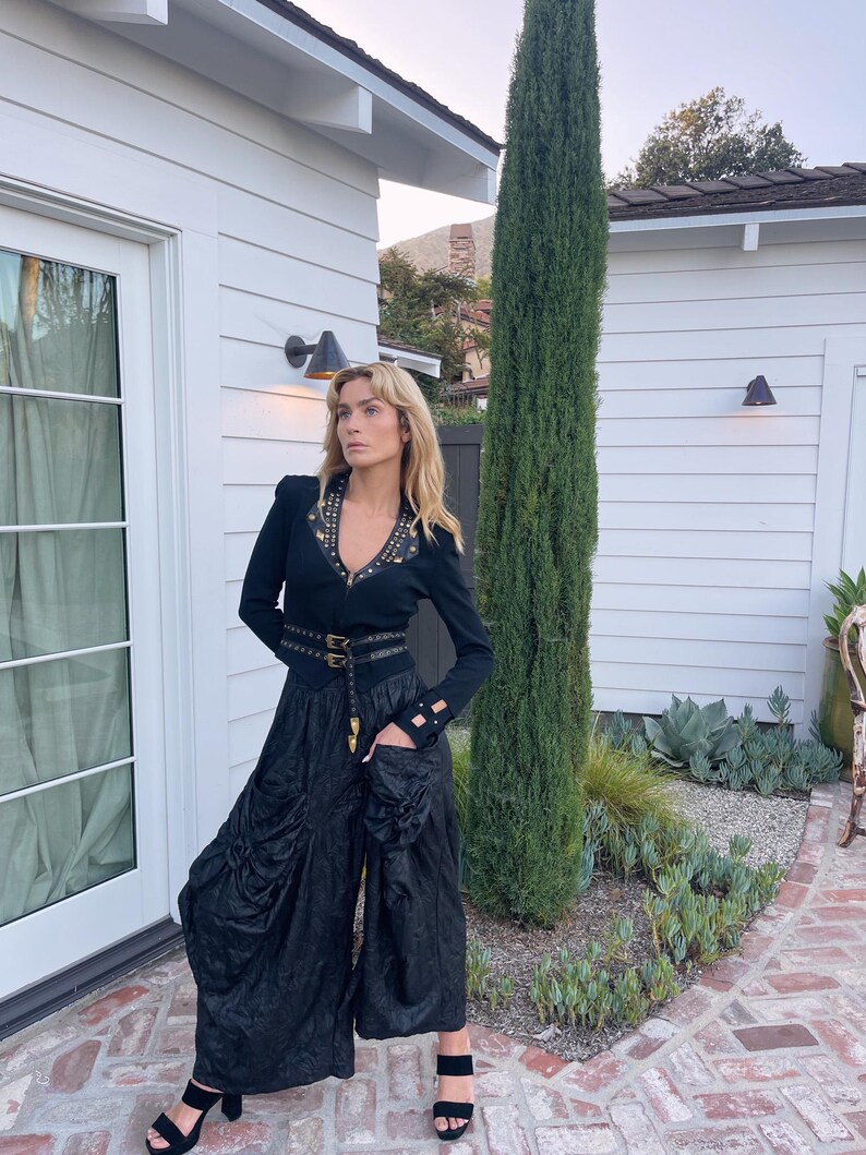 May include: A black long-sleeved top with gold-colored stud accents and a matching belt. The outfit includes wide-leg black pants and black heeled sandals. A tall, green tree stands in the background, with a white building and brick pathway.