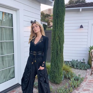 May include: A black long-sleeved top with gold-colored embellishments and a matching belt, paired with black, wide-leg pants. The outfit is accessorized with black sandals. The background features a white building and greenery.