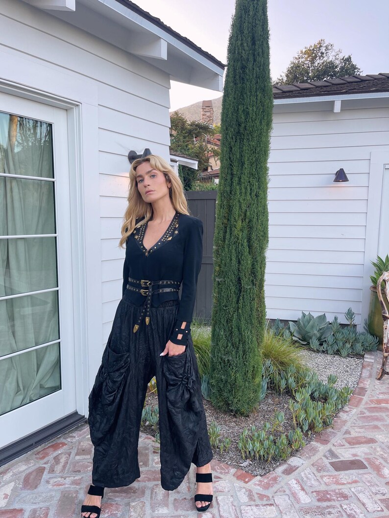 May include: A black long-sleeved top with gold-colored embellishments and a matching belt, paired with black, wide-leg pants. The outfit is accessorized with black sandals. The background features a white building and greenery.