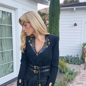 May include: Black jacket with gold-colored stud accents and a matching belt with double buckles. The jacket has a zipper closure and cut-out details on the sleeves. The outfit is completed with a black skirt.