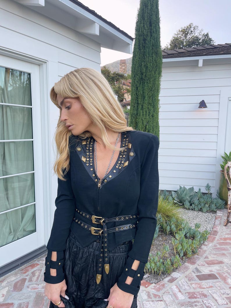 May include: Black jacket with gold-colored stud accents and a matching belt with double buckles. The jacket has a zipper closure and cut-out details on the sleeves. The outfit is completed with a black skirt.