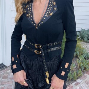 May include: Black jacket with gold-colored stud and star accents, a leather-like collar, and a matching belt with gold buckles. The jacket has cut-out details on the sleeves and a zipper closure. The outfit is completed with black pants.