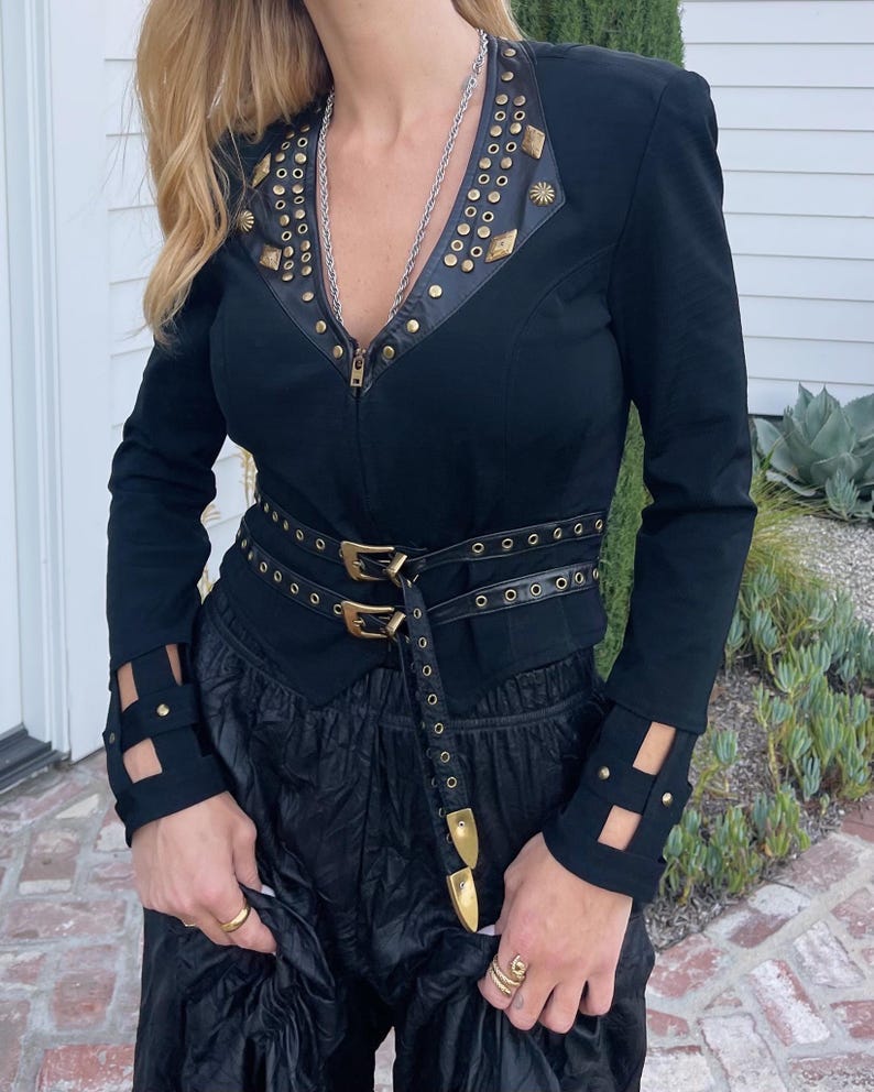 May include: Black jacket with gold-colored stud and star accents, a leather-like collar, and a matching belt with gold buckles. The jacket has cut-out details on the sleeves and a zipper closure. The outfit is completed with black pants.