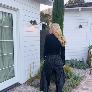 May include: Black long-sleeve top and wide-leg pants. The pants have a textured appearance. The person is wearing black platform sandals. The background includes a white building, a tall green tree, and a brick patio.