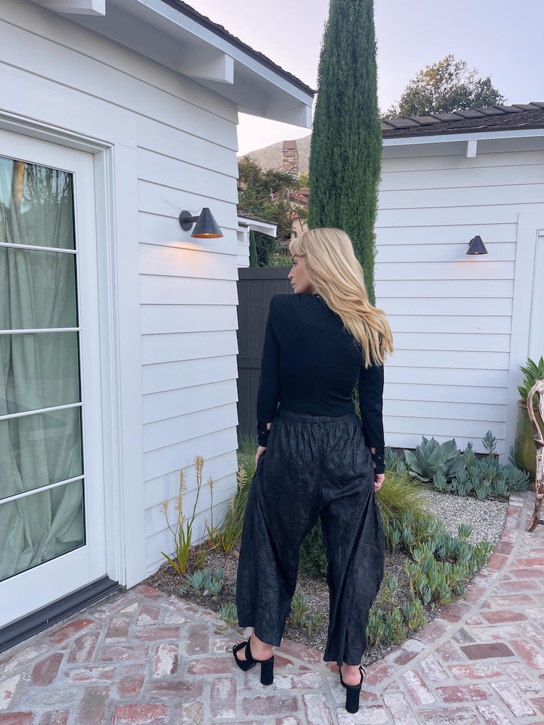 May include: Black long-sleeve top and wide-leg pants. The pants have a textured appearance. The person is wearing black platform sandals. The background includes a white building, a tall green tree, and a brick patio.