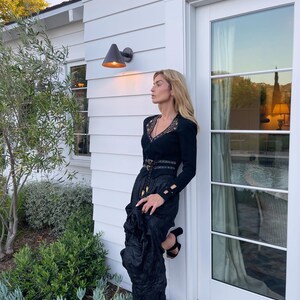 May include: A black long-sleeved top with lace detailing and a matching black skirt. The outfit is accessorized with a belt and black heeled sandals. The woman is leaning against a white wall next to a door.