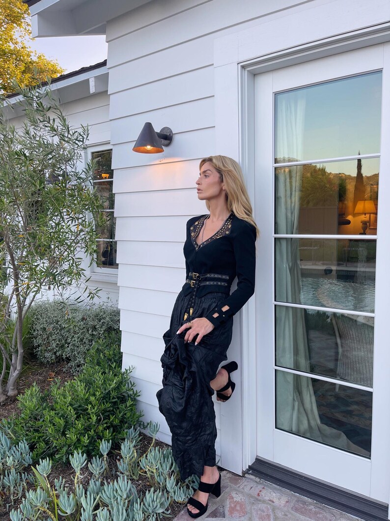 May include: A black long-sleeved top with lace detailing and a matching black skirt. The outfit is accessorized with a belt and black heeled sandals. The woman is leaning against a white wall next to a door.