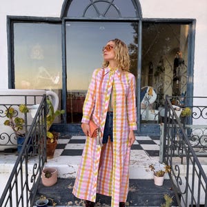 May include: A long-sleeved, floor-length button-down shirt dress featuring a vibrant pink, orange, and yellow plaid pattern. The dress is worn open over dark blue denim shorts, paired with dark red lace-up boots, a peach clutch, and sunglasses.