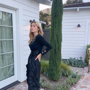 May include: A black long-sleeved top with gold embellishments and a matching black skirt. The skirt has a gathered, textured design. The outfit is worn outdoors near a white building and a tall, green tree.