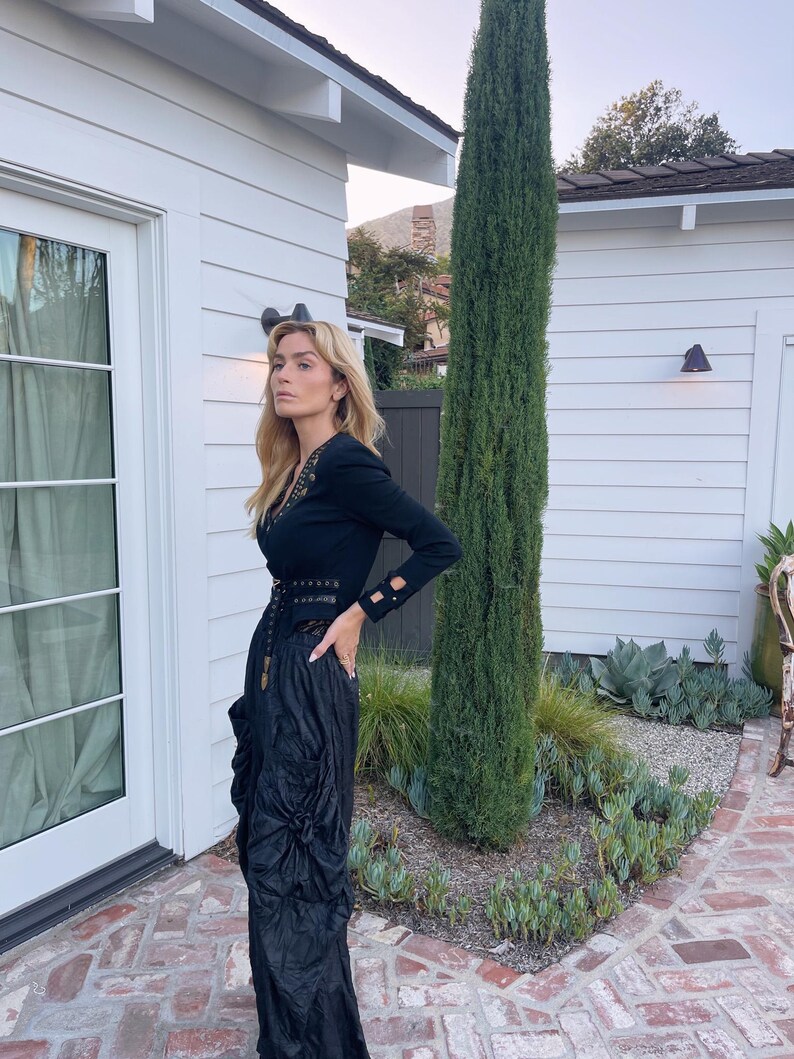 May include: A black long-sleeved top with gold embellishments and a matching black skirt. The skirt has a gathered, textured design. The outfit is worn outdoors near a white building and a tall, green tree.