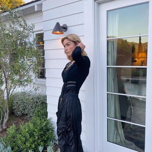 May include: A woman wearing a black outfit, including a long-sleeved top with a lace neckline and a high-waisted skirt with a draped design. The outfit is paired with black heeled sandals. The background features a white house with a door and a garden.