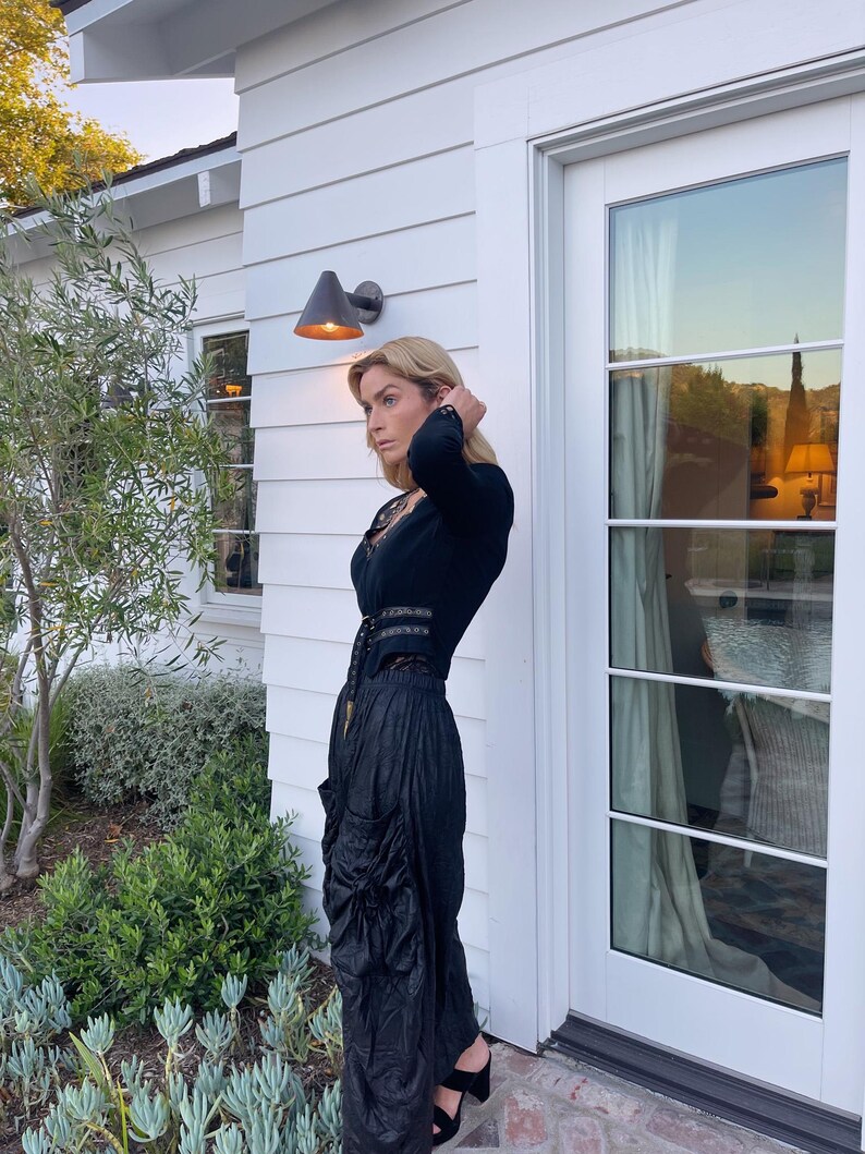 May include: A woman wearing a black outfit, including a long-sleeved top with a lace neckline and a high-waisted skirt with a draped design. The outfit is paired with black heeled sandals. The background features a white house with a door and a garden.