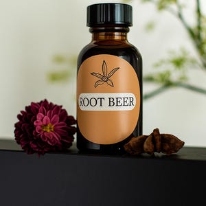 以下が含まれることがあります： 黒いキャップと「ROOT BEER」と書かれたラベルが付いた茶色のガラス瓶。ラベルには花のイラストがあります。濃い赤色の花と茶色のスパイスがボトルの横にあります。ボトルは黒い表面にあります。