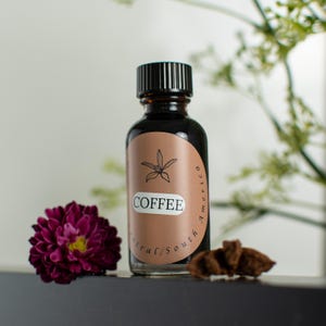 Könnte beinhalten: Eine dunkle Glasflasche mit schwarzem Deckel und einem Etikett mit der Aufschrift "COFFEE" mit einer Blumenillustration. Das Etikett ist braun mit den Worten "Central/South America" in geschwungener Schrift. Eine dunkelviolette Blume und ein kleines braunes Objekt befinden sich neben der Flasche.