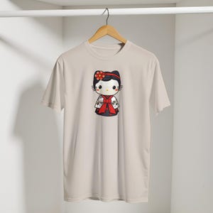 May include: A light beige t-shirt featuring a cartoon character in a traditional Japanese kimono. The character has a black bow, a red flower, and a red bow. The kimono is black and white with red accents. The shirt is hanging on a wooden hanger.
