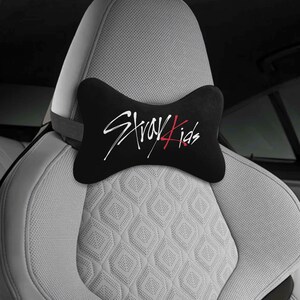 May include: Black car headrest pillow with the text "Stray Kids" in white and red script. The pillow is shaped like a bow tie and is placed on a light gray car seat with a diamond pattern.