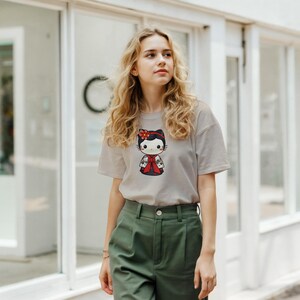 May include: A light gray t-shirt featuring a cartoon cat character in a traditional Japanese outfit. The cat has a red flower in its hair and is wearing a red and black kimono. The shirt is paired with olive green pants.