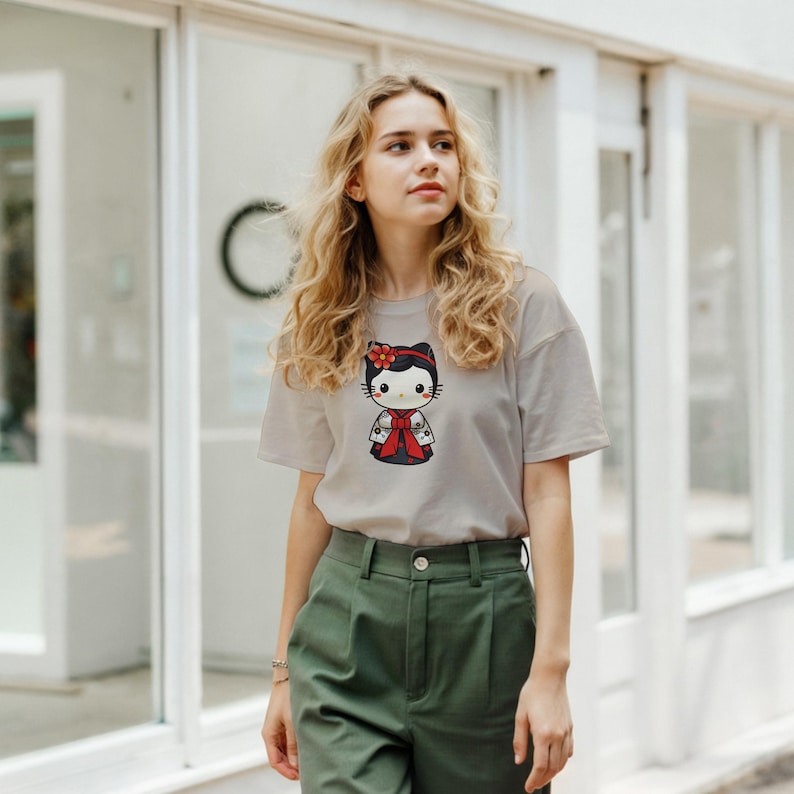 May include: A light gray t-shirt featuring a cartoon cat character in a traditional Japanese outfit. The cat has a red flower in its hair and is wearing a red and black kimono. The shirt is paired with olive green pants.