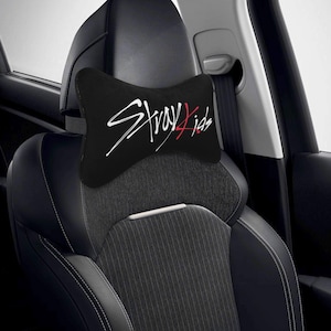 May include: Black car headrest pillow with the text "Stray Kids" in white and red script. The pillow is shaped like a bow tie and is placed on a car seat. The car seat is black and gray.