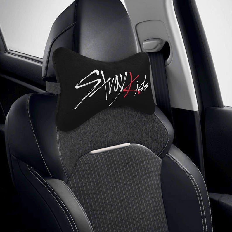 May include: Black car headrest pillow with the text "Stray Kids" in white and red script. The pillow is shaped like a bow tie and is placed on a car seat. The car seat is black and gray.
