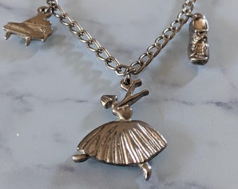 May include: Silver-toned charm bracelet with a ballerina, piano, and ballet slipper charms. The ballerina is in a graceful pose, and the piano has detailed keys. The ballet slipper charm has intricate lace details.