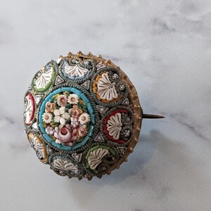 Pode incluir: Broche redondo de mosaico antigo com um design floral. O broche apresenta um grupo central de flores rosa e brancas rodeado por uma borda turquesa. O anel externo tem sec&ccedil;&otilde;es alternadas de azulejos de mosaico coloridos e formas brancas em forma de folha.