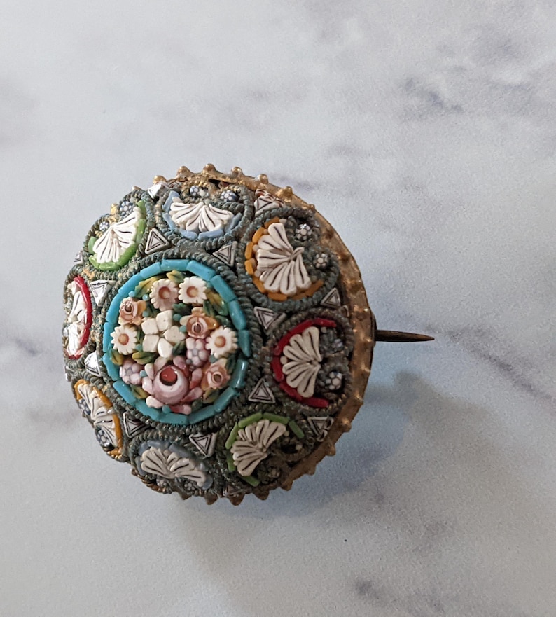 Pode incluir: Broche redondo de mosaico antigo com um design floral. O broche apresenta um grupo central de flores rosa e brancas rodeado por uma borda turquesa. O anel externo tem sec&ccedil;&otilde;es alternadas de azulejos de mosaico coloridos e formas brancas em forma de folha.