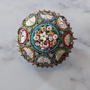Pode incluir: Broche redondo de mosaico com um design floral. O broche apresenta um grupo central de flores brancas e rosas rodeado por uma borda turquesa. A borda externa tem uma moldura de metal dourado com detalhes de mosaico coloridos.