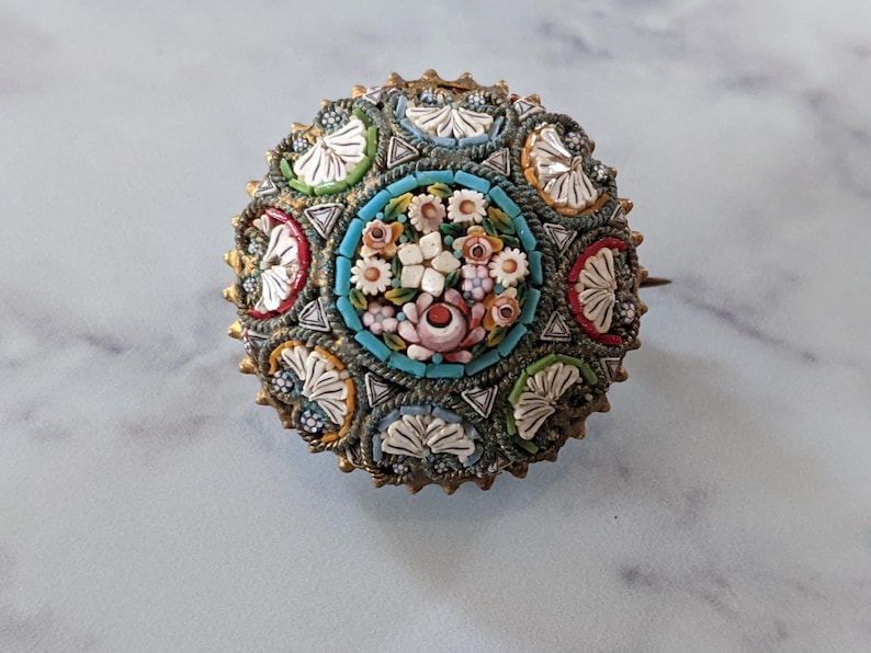 Pode incluir: Broche redondo de mosaico com um design floral. O broche apresenta um grupo central de flores brancas e rosas rodeado por uma borda turquesa. A borda externa tem uma moldura de metal dourado com detalhes de mosaico coloridos.