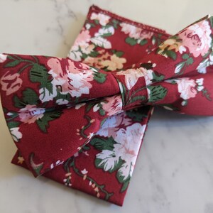 May include: A burgundy bow tie and matching pocket square set. The fabric features a floral pattern with white, pink, and green flowers. The bow tie is pre-tied, and the pocket square is folded neatly.