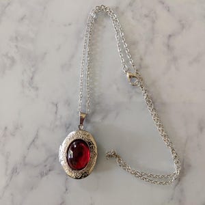 Könnte beinhalten: Silberfarbene ovale Medaillon-Halskette mit einem roten Edelstein in der Mitte. Das Medaillon hat ein dekoratives Blumendesign an den Rändern und hängt an einer zarten Kette. Die Halskette wird auf einer Marmoroberfläche präsentiert.