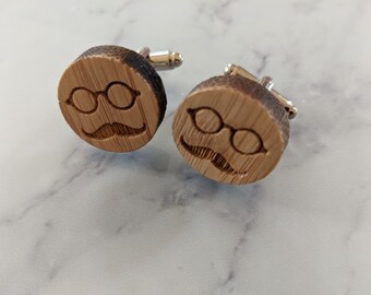 May include: A pair of wooden cufflinks with a silver-colored backing. Each cufflink features a carved design of eyeglasses and a mustache. The cufflinks are round and made of light brown wood.
