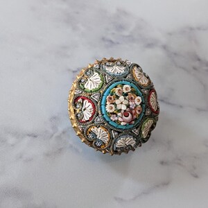 Pode incluir: Um objeto decorativo redondo, estilo mosaico, com um design floral. O centro apresenta um buqu&ecirc; de flores em rosa, branco e p&ecirc;ssego, rodeado por uma borda turquesa. A borda externa tem uma moldura dourada e padr&otilde;es de mosaico.
