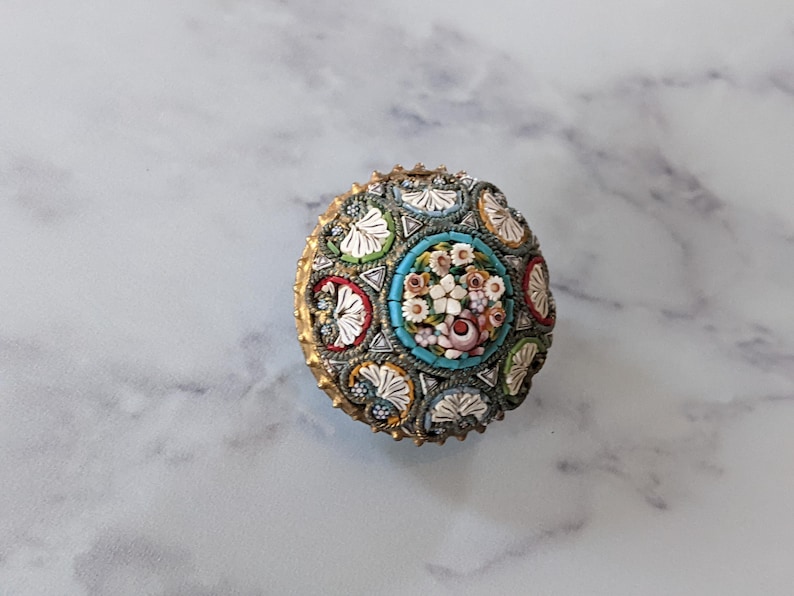 Pode incluir: Um objeto decorativo redondo, estilo mosaico, com um design floral. O centro apresenta um buqu&ecirc; de flores em rosa, branco e p&ecirc;ssego, rodeado por uma borda turquesa. A borda externa tem uma moldura dourada e padr&otilde;es de mosaico.