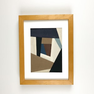 Könnte beinhalten: Abstrakte geometrische Kunst in einem hellen Holzrahmen. Das Kunstwerk zeigt eine Collage aus Stoffstücken in verschiedenen Formen und Farben, darunter Marineblau, Braun, Beige und Schwarz. Das Kunstwerk ist auf einem weißen Passepartout montiert.