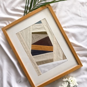 Könnte beinhalten: Ein gerahmtes abstraktes Kunstwerk mit einem hellen Holzrahmen. Das Kunstwerk zeigt ein geometrisches Design mit verschiedenen Stoffstreifen in neutralen Tönen, darunter Beige, Braun und Blau. Ein grünes Palmenblatt und weiße Blumen sind im Hintergrund.