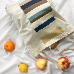 Könnte beinhalten: Eine beigefarbene Tragetasche mit einem farbenfrohen Streifendesign, die ein Brot enthält. Die Tasche ist von einer Orange, einer Zitrone und zwei Äpfeln umgeben. Die Tasche hat lange Träger.