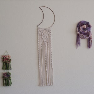 May include: A white macrame wall hanging with a crescent moon shape at the top, hanging on a white wall. There are three smaller macrame wall hangings with floral designs below the larger one.