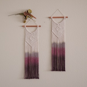May include: Two macrame wall hangings with a geometric design and a gradient of pink, purple, and gray colors. The hangings are made with natural cotton cord and have a wooden dowel at the top.