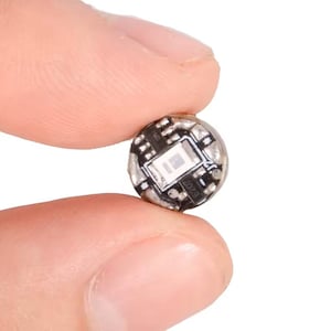May include: A small, circular electronic component featuring a central square LED light mounted on a black circuit board base. The component displays visible metallic soldering points and tiny printed numerical markings on its surface.