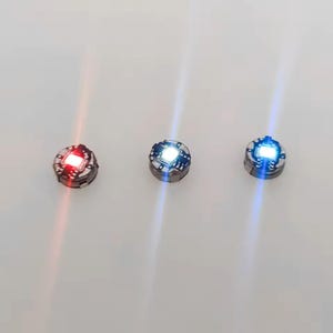 May include: Three small circular electronic components with integrated LED lights. The first emits a bright red glow, the second a neutral white light, and the third a vibrant blue light. Each unit features a dark circuit board base.