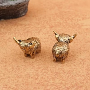 Könnte beinhalten: Zwei kleine, goldfarbene Metallfiguren in Form von Hochlandrindern. Jedes Miniaturtier weist detaillierte, texturierte Fellmuster, geschwungene Hörner und eine kompakte, robuste Haltung auf, die das zottelige Aussehen der Rasse einfängt.