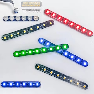 May include: Multiple linear LED light strips in red, green, blue, and warm white, each featuring a series of small surface-mounted diodes on a dark circuit board. Text on the boards includes "7986769A_Y33_251110" and "CR927". A strip of six 3V button batteries is shown with tweezers.
