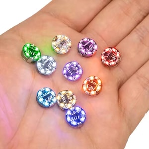 May include: A collection of ten small, circular LED light inserts, each featuring a central electronic component and emitting a distinct glow in colors including green, white, purple, red, orange, and blue, arranged in a scattered pattern.