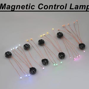 Könnte beinhalten: Ein Set aus Magnetic Control Lamp Komponenten mit kleinen schwarzen runden Basen, die mit dünnen Kupferdrähten verbunden sind, an denen winzige leuchtende LED-Lichter in verschiedenen Farben wie Weiß, Gelb, Grün, Pink und Blau befestigt sind.