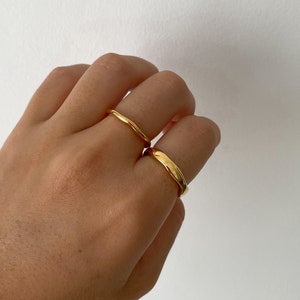 May include: Two gold rings on a hand. Both rings are simple and have a smooth, rounded surface. The ring on the index finger is thinner than the ring on the middle finger.